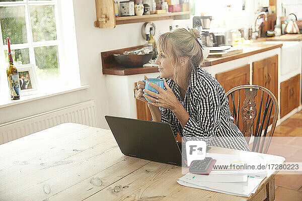 Seniorin hält Kaffeetasse und schaut weg,  während sie mit Laptop auf dem Tisch in der Küche zu Hause sitzt
