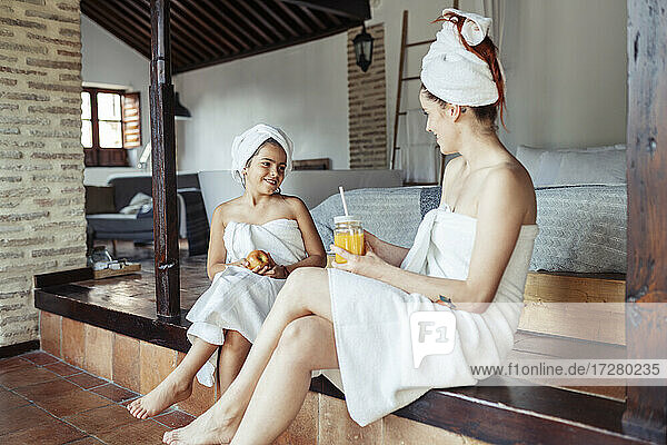 Lächelnde Frau und Mädchen in Handtuch mit Obst und Saft beim Sitzen zu Hause