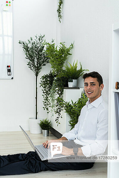 Lächelnder männlicher Unternehmer mit Laptop  der bei Pflanzen im Büro auf dem Boden sitzt