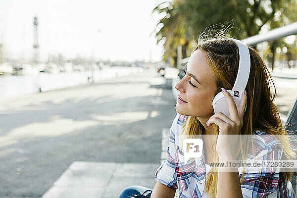 Young woman listening music through headphone sitting on footpath during sunny day