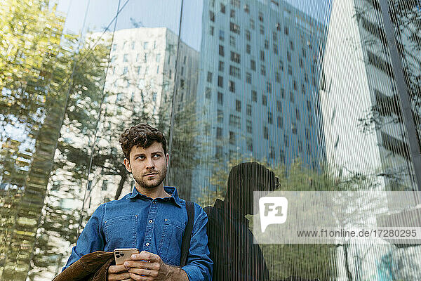 Thoughtful young businessman leaning on glass wall of modern office building