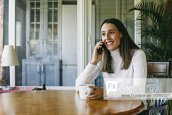 Junge Frau mit Kaffeetasse  die zu Hause sitzt und mit ihrem Handy telefoniert