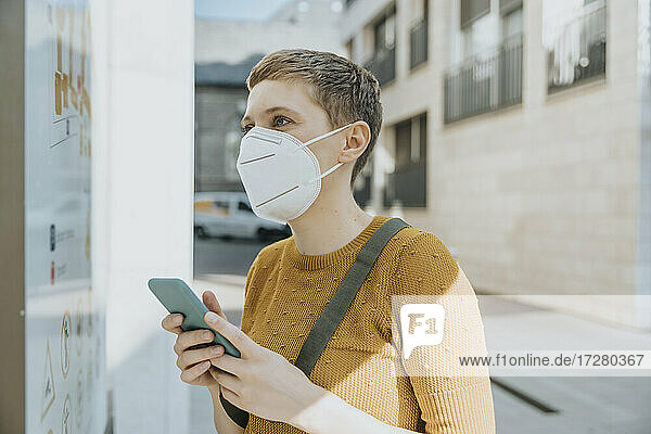 Frau mit Gesichtsschutzmaske  die an einem sonnigen Tag in der Stadt steht und eine Karte liest  während sie ihr Smartphone in der Hand hält