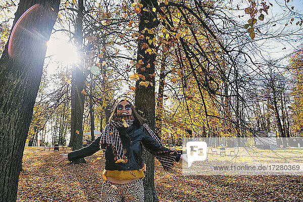 Glückliche Frau steht mit ausgestreckten Armen unter fallenden trockenen Herbstblättern vor Bäumen im Park