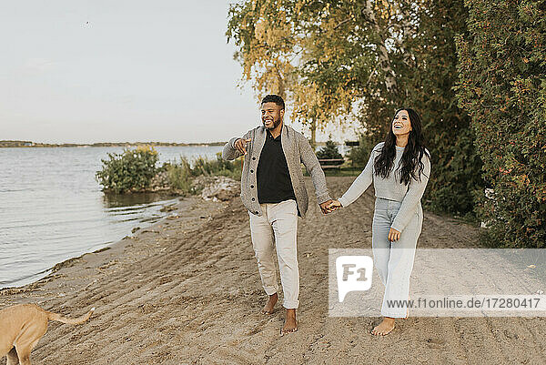 Lächelnder Freund und Freundin halten sich beim Spaziergang mit Hund am See an den Händen