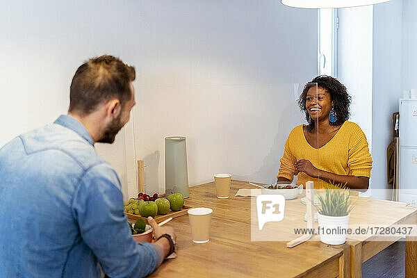 Lächelnde Frau beim Essen mit einem Mitarbeiter  der an einem Tisch mit Bildschirmteilung im Büro sitzt