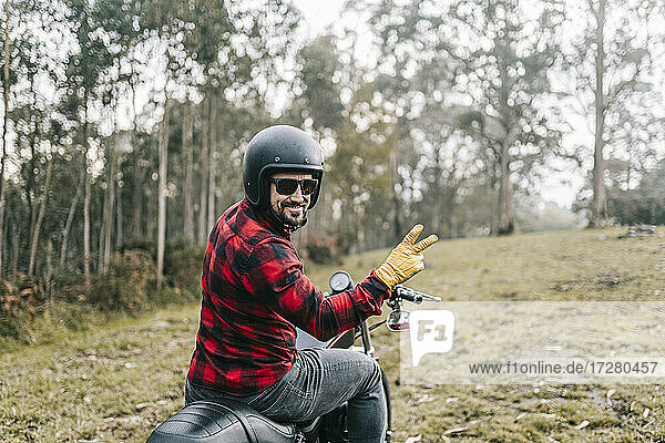 Lächelnder männlicher Biker  der auf einem Motorrad den Wald erkundet  gestikuliert