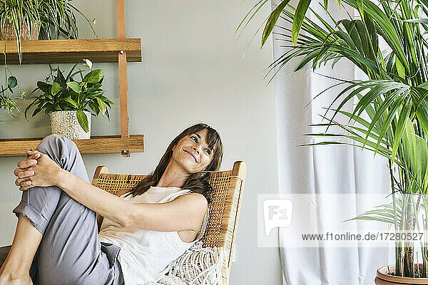 Lächelnde Frau  die ihr Knie umarmt  während sie sich auf einem Stuhl zu Hause entspannt