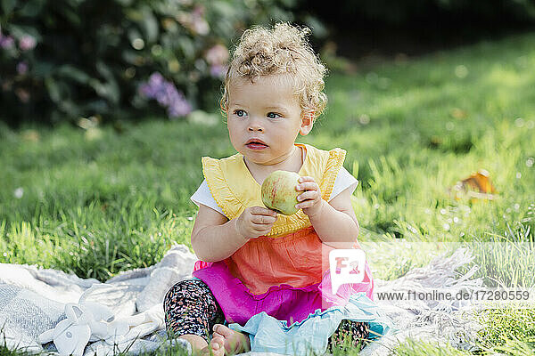 Cute baby girl holding fruit while sitting on grass at park