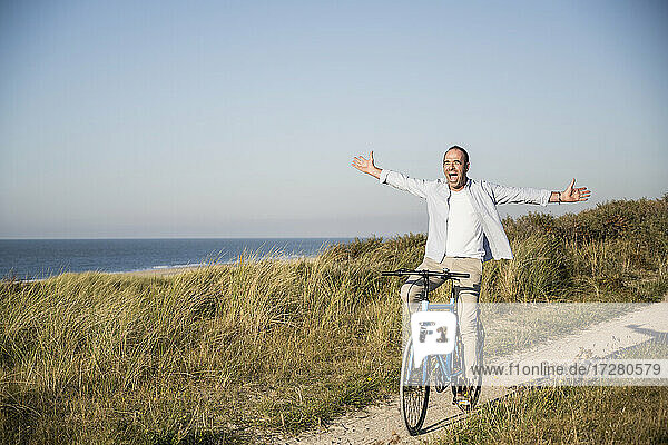 Happy mature man with arms outstretched riding bicycle at beach against clear sky