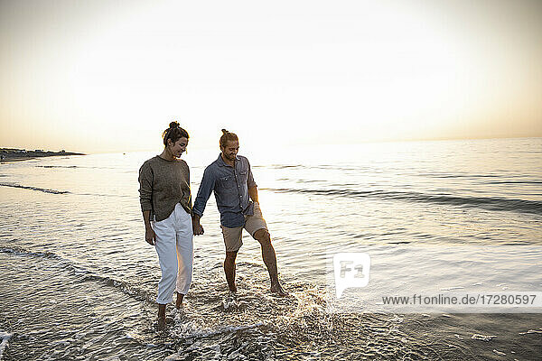 Happy young couple holding hands while walking on shore at beach during sunset