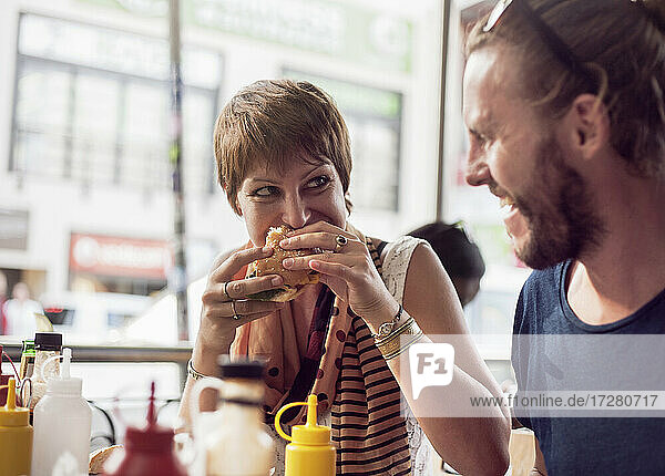 Junge Frau isst Burger  während sie mit einem erwachsenen Mann in einem Cafe sitzt