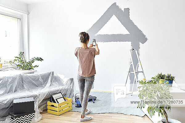 Junge Frau  die mit ihrem Handy ein Foto von einem an die Wand gemalten Haus macht  während sie zu Hause steht