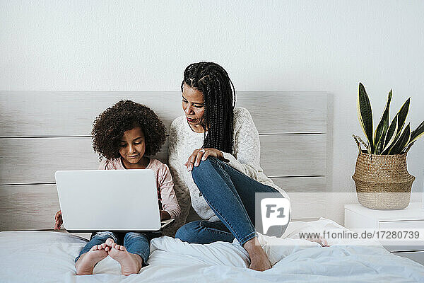 Tochter benutzt Laptop  während die Mutter neben ihr auf dem Bett im Schlafzimmer sitzt