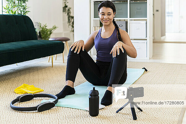 Smiling young woman video recording while sitting on exercise mat at home