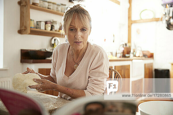 Senior woman kneading while reading recipe book sitting in kitchen
