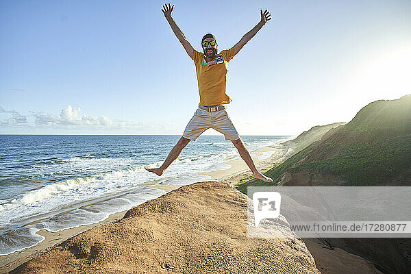 Fröhlicher Mann  der auf eine Felsformation am Strand springt  gegen blauen Himmel
