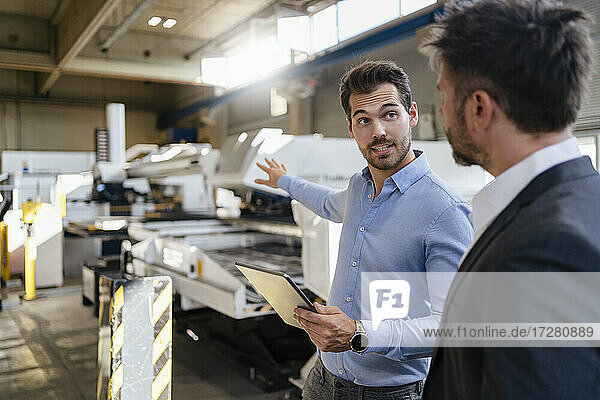 Young businessman with digital tablet gesturing toward equipment while standing by colleague at factory