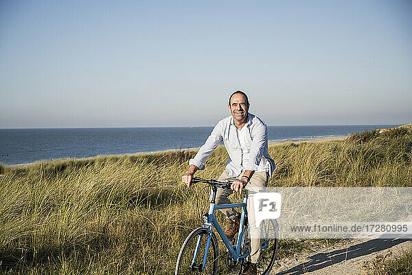 Smiling mature man riding bicycle at beach against clear sky