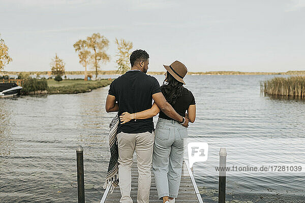 Boyfriend and girlfriend with arm around walking on pier against lake