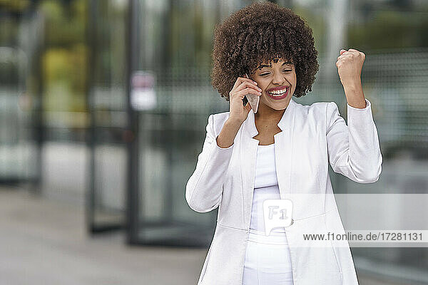 Glückliche Geschäftsfrau  die eine gewinnende Geste zeigt  während sie mit einem Mobiltelefon gegen ein Gebäude spricht