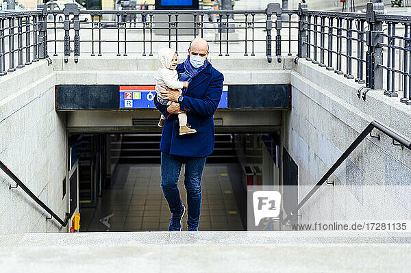 Man with face mask carrying baby while walking on staircase in city