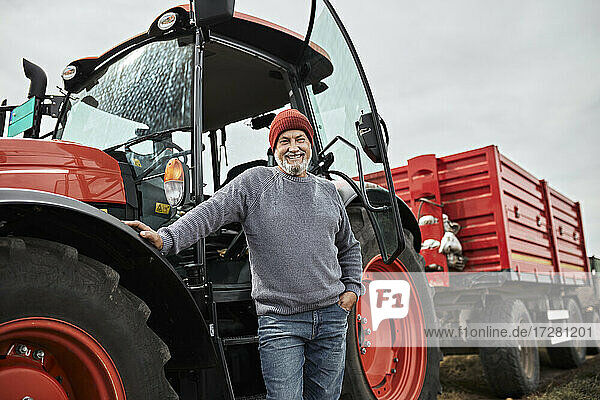 Lächelnder reifer Landwirt steht mit der Hand in der Tasche vor einem roten Traktor auf einem Bauernhof