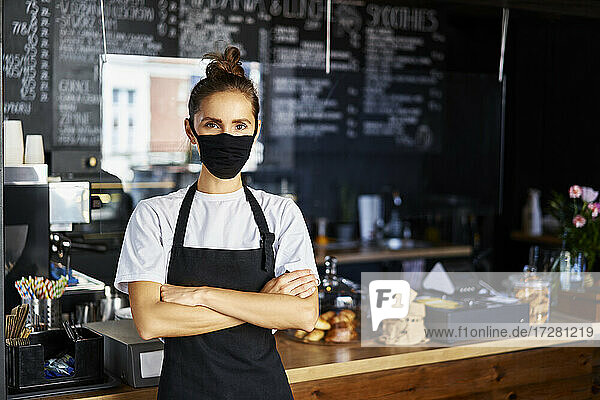 Portrait of waitress in protective face mask with arms crossed standing at cafe