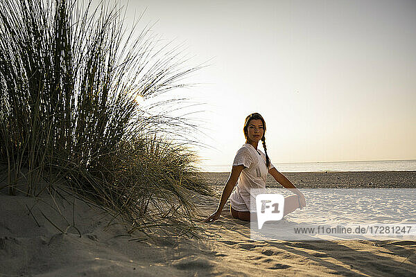 Schöne junge Frau übt Yoga  während sie am Strand gegen den klaren Himmel bei Sonnenuntergang sitzt