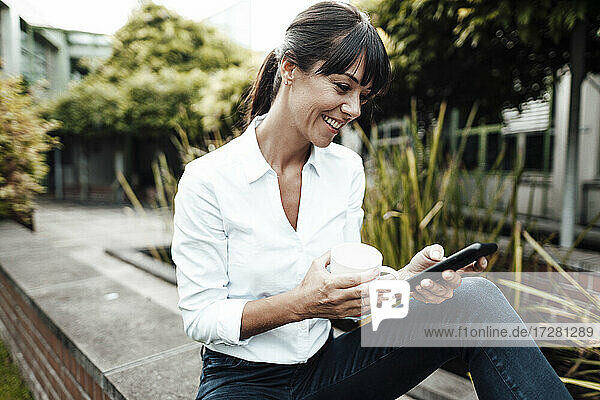 Lächelnde weibliche Fachkraft  die eine Kaffeetasse hält  während sie in der Pause ihr Smartphone benutzt