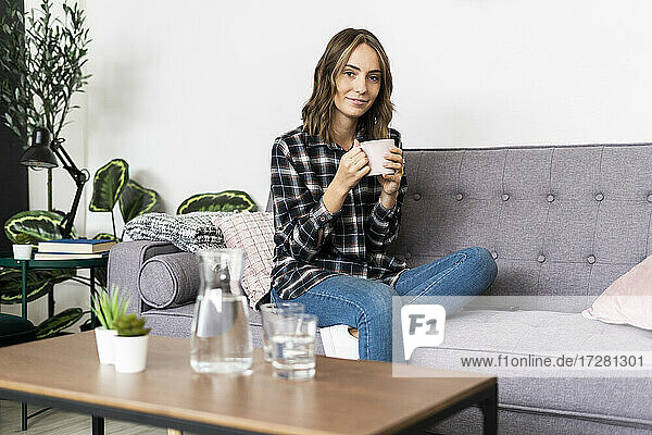 Junge Frau trinkt Kaffee  während sie zu Hause auf dem Sofa sitzt