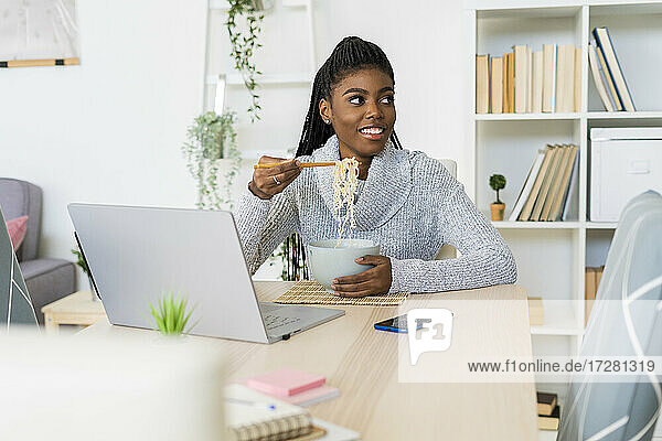 Lächelnde junge Frau  die wegschaut  während sie Nudeln isst und zu Hause sitzt