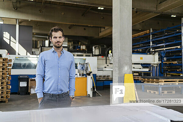 Confident businessman with hands in pockets standing by table at factory