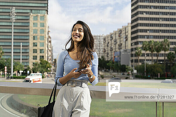 Smiling businesswoman holding smart phone while leaning on railing with city in background