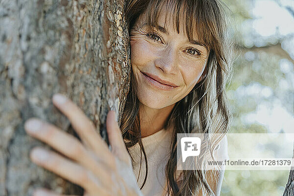 Lächelnde reife Frau lehnt an einer Kiefer in einem öffentlichen Park