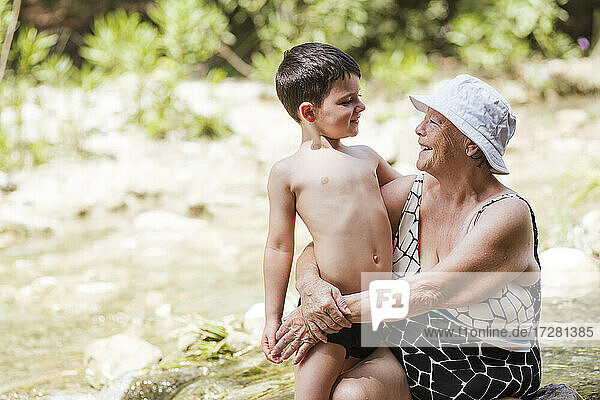 Grandson and grandmother smiling while sitting in river at forest