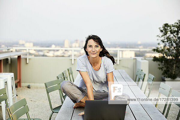 Lächelnde reife Frau im Schneidersitz mit Laptop auf der Terrasse eines Gebäudes sitzend