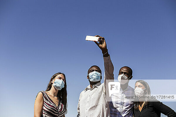 Business-Profis nehmen Selfie mit schützenden Gesichtsmasken gegen klaren blauen Himmel an einem sonnigen Tag