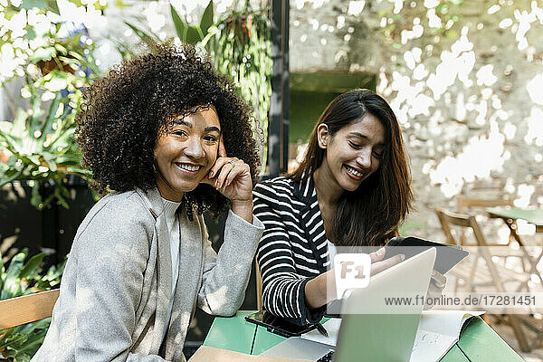 Smiling female coworkers smiling using laptop and digital tablet sitting in cafe
