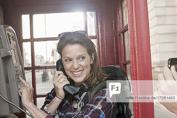 Smiling woman holding telephone receiver while looking at friend photographing through smart phone