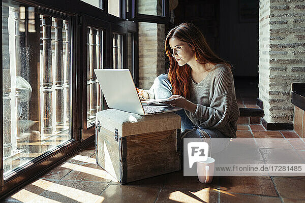 Rothaarige Frau arbeitet am Laptop  während sie zu Hause auf dem Boden sitzt