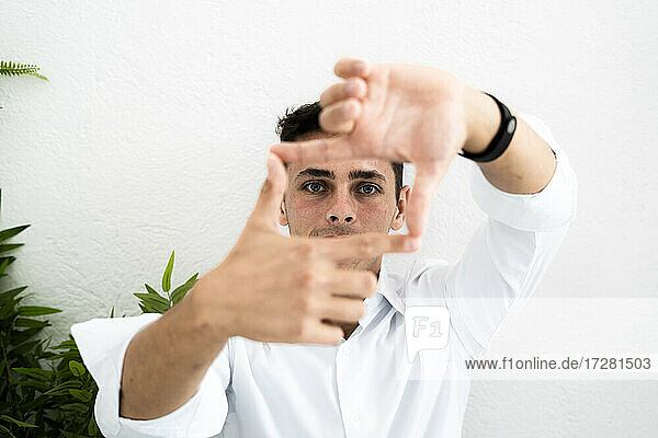 Male entrepreneur making square shape from fingers and thumbs against white wall at office