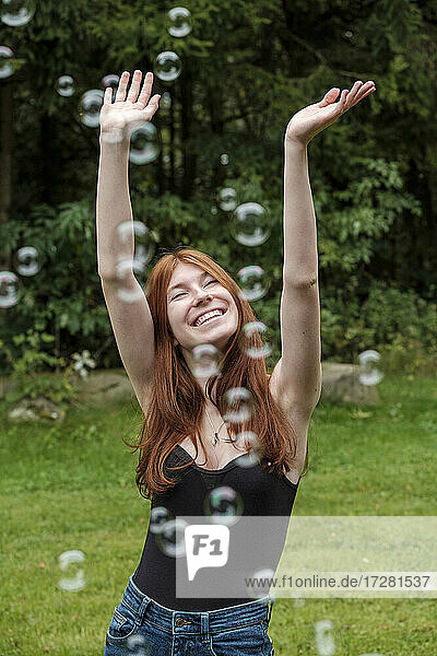 Happy woman with arms raised playing amidst soap bubbles in yard