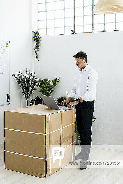 Junger Geschäftsmann  der einen Laptop benutzt  während er an einem kreativen Arbeitsplatz neben einer Pappschachtel steht