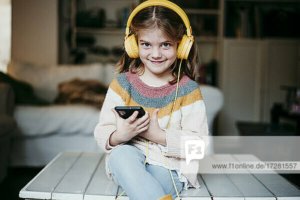 Lächelndes Mädchen mit Kopfhörern  das ein Mobiltelefon benutzt  während es zu Hause auf dem Tisch sitzt