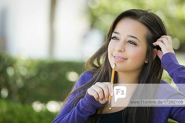 Attractive pensive mixed-race female student with pencil sitting on campus bench