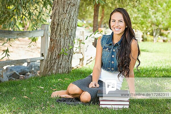 Glückliche gemischtrassige junge Studentin mit Büchern und Computer-Tablet draußen sitzen im Gras