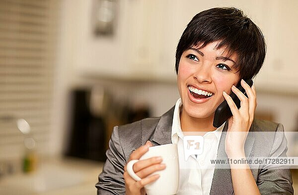 Pretty smiling multiethnic woman with coffee and talking on a cell phone in her kitchen