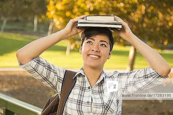 Porträt einer hübschen gemischtrassigen jungen Frau  die an einem sonnigen Nachmittag im Park Bücher auf dem Kopf hält