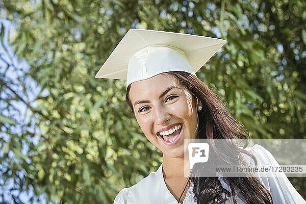 Attraktives lächelndes gemischtrassiges Mädchen  das draußen mit Kappe und Talar seinen Abschluß feiert